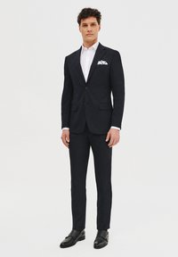 Granatowy garnitur o slim fit, jednorzędowa marynarka z jednym guzikiem, dopasowane spodnie, biała koszula i wzorzysta poszetka. Czarne skórzane buty dopełniają całość.