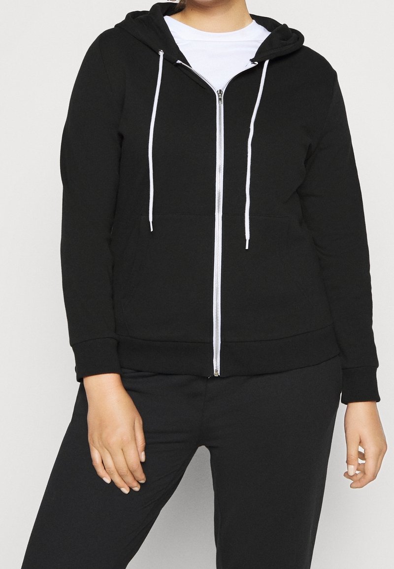 Personne portant un sweat à capuche noir zippé avec des cordons blancs sur une chemise blanche, associé à un pantalon noir, debout devant un fond uni.