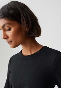 Frau mit kurzem dunklem Haar, die ein schlichtes schwarzes Rundhals-T-Shirt trägt und nach unten schaut, vor einem schlichten hellen Hintergrund.