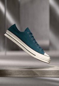 Teal plátené tenisky s nízkym strihom, bielym gumovým špičkou a čiernym pruhom pozdĺž podrážky. Obsahuje šesť dierok a textúrovaný povrch.
