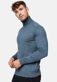 Jersey de cuello alto azul claro hecho de una suave tela de punto. Presenta puños y dobladillo acanalados, corte ajustado y un sutil logo en la parte inferior.