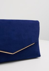 Clutch de ante navy con solapa triangular, que presenta un detalle de metal dorado a lo largo del borde. Textura suave con sutiles detalles de costura.