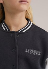 Temno siva univerzitetna jakna z belimi črtastimi rebernimi ovratnikom, s pritisnimi gumbi in vezenim napisom "AU REVOIR" spredaj.