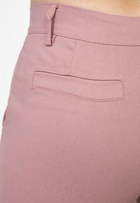 Pantalons rose clair en tissu texturé, avec une poche plate au dos et une boucle sécurisée à la taille. Des détails de couture lisse sont visibles.