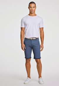 Pantaloni corti in cotone blu con orli arrotolati, abbinati a una t-shirt bianca a girocollo e sneakers bianche. La cintura presenta una texture intrecciata e un accento marrone.