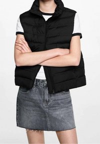 Svart vadderad väst med hög krage och dragkedja framtill, bärs över en vit randig skjorta. Matchas med en grå jeanskjol.