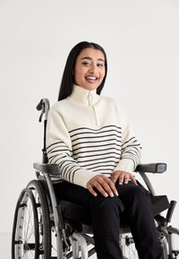 Maglione lavorato a maglia con colletto alto, colore crema con strisce orizzontali nere, dotato di una zip parziale frontale e polsini a costine, indossato da una persona su una sedia a rotelle.