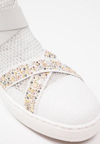 Baskets blanches en tissu mesh avec des brides croisées décorées de clous multicolores en tons or, argent et rose.