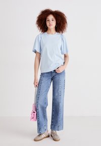 T-shirt di cotone azzurro chiaro con maniche arricciate, abbinata a jeans in denim a gamba larga, sneakers grigie e una piccola borsa rosa.
