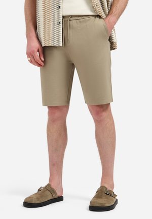 Man draagt beige shorts met trekkoord, een overhemd met patroon en bruine instapschoenen met gespen, staand met handen in de zakken.