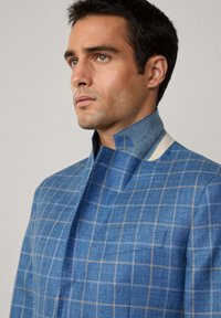 Giacca a scacchi blu con un alto colletto e cuciture a contrasto. Il tessuto ha una finitura testurizzata con accenti dorati a griglia.