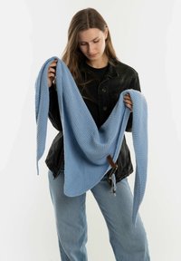 Lichtblauwe gebreide sjaal met een gemarcreerd patroon, vastgehouden door een model in een zwarte jas, met een decoratief label en franjes aan de randen.