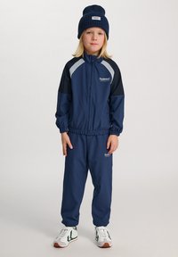 Kind trägt eine marineblaue Sport-Trainingsanzug mit weißem Logo, passender Strickmütze und weißen Turnschuhen, steht vor einem einfarbigen Hintergrund.