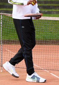 Pantaloni atletici negri cu dungi laterale albastre, pantofi albi și gri cu un accent verde neon și o jachetă albă cu fermoar, ținând o rachetă de tenis și o minge.