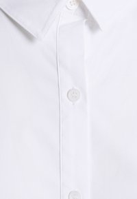 Chemise blanche à col pointu avec des boutons en plastique sur la patte de boutonnage avant.
