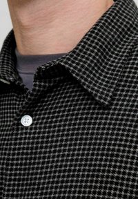 Camisa de cuadros en negro y gris con cuello y un botón blanco. La tela presenta un patrón texturizado con un sutil tejido.