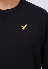 T-shirt en coton noir avec un petit logo d'oiseau en or brodé sur la poitrine. Encolure ronde classique avec un détail de couture texturé.