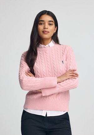 Suéter de punto en color rosa claro con puños y dobladillo acanalados, que presenta un pequeño logo negro. Llevado sobre una camisa blanca de cuello, con pantalones oscuros visibles.