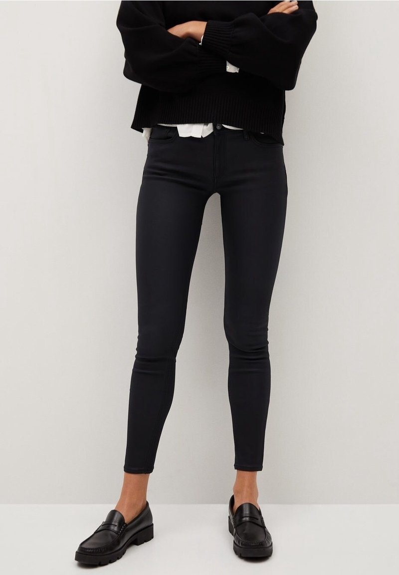 Femme portant un jean skinny noir, des mocassins noirs, un pull noir et une chemise blanche, debout les bras croisés.