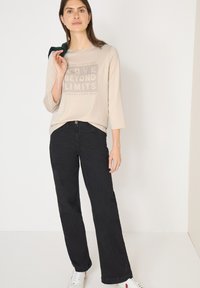 Beżowa bluzka z długim rękawem z nadrukowanym tekstem, zestawiona z czarnymi spodniami o szerokich nogawkach. Modelka ma na sobie białe trampki i trzyma zielony sweter.