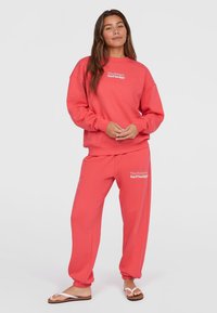 Korálová súprava loungewear obsahujúca mikinu s okrúhlym výstrihom a tepláky s elastickými manžetami. Obe položky majú biele textové logo na hrudi a stehne.