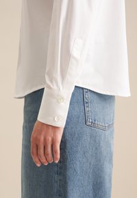 Camicia bianca con colletto, maniche lunghe e polsini abbottonati, abbinata a una gonna di jeans a vita alta, realizzata in un tessuto testurizzato e di colore blu.