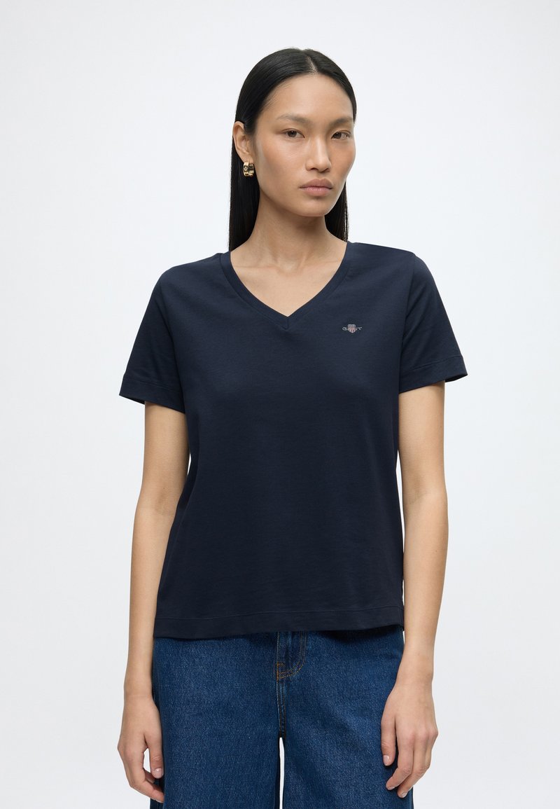 Femme aux longs cheveux noirs portant des boucles d'oreilles anneaux en or, un t-shirt bleu marine à col en V et un jean bleu, debout devant un fond blanc.