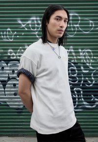 Fehér texturált ing kék kockás mandzsettával, fekete rövidnadrággal párosítva. Ezüst lánc nyaklánccal kiegészítve, zöld háttér előtt.