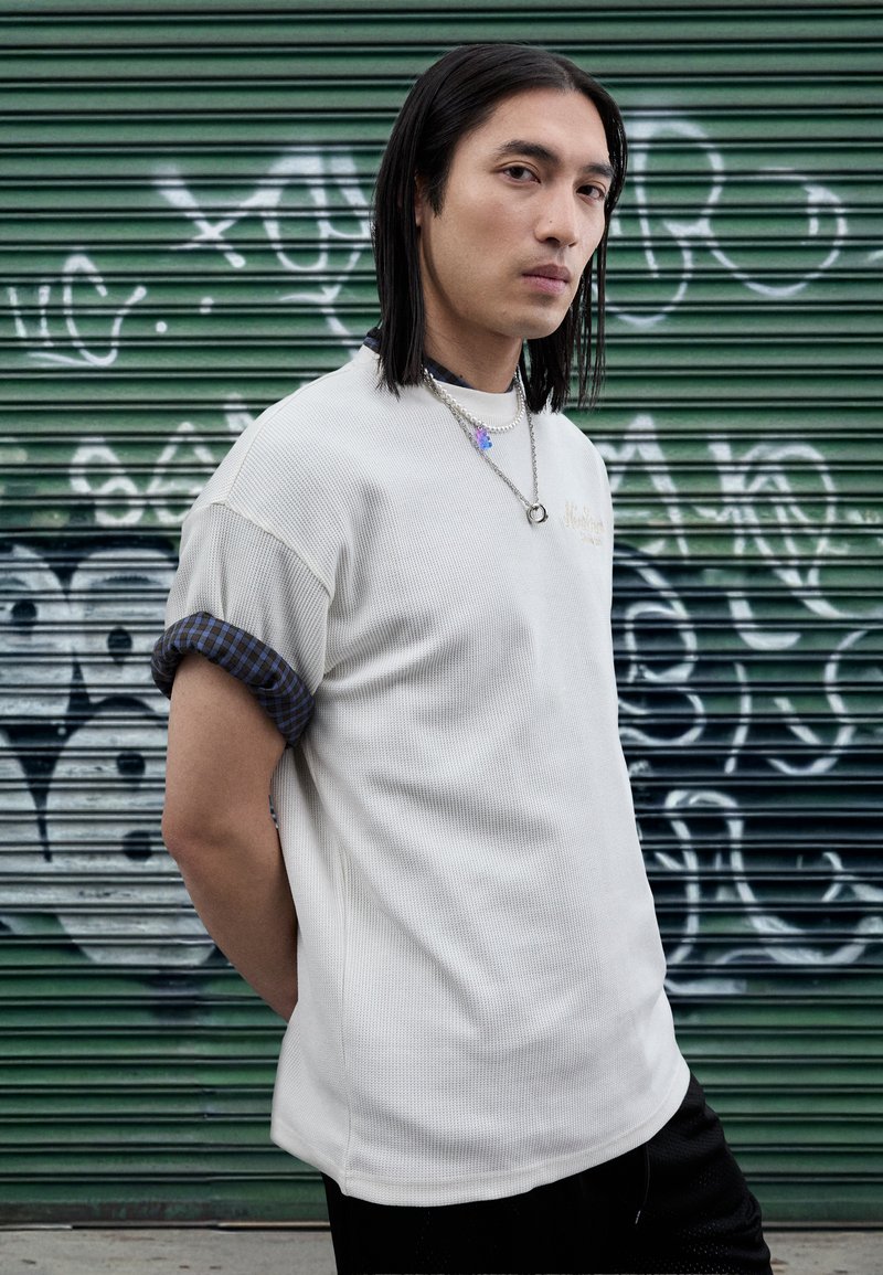 Fehér texturált ing kék kockás mandzsettával, fekete rövidnadrággal párosítva. Ezüst lánc nyaklánccal kiegészítve, zöld háttér előtt.