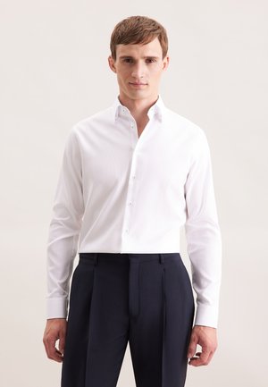 Man in een witte, ingebuttonde shirt met lange mouwen, gestoken in hoge donkerblauwe geplooide broek, staande tegen een effen achtergrond.
