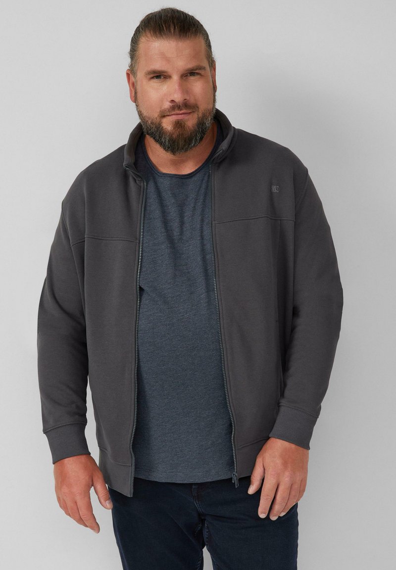 Giacca zip grigia scura con colletto alto, caratterizzata da una superficie liscia e un design minimalista; indossata sopra una maglietta blu a maniche corte.