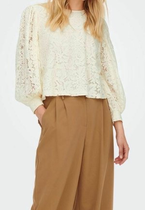 Kobieta w kremowej koronkowej bluzce z długim rękawem, bufkami na rękawach oraz w brązowych szerokich spodniach z wysokim stanem.