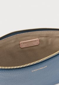 Bolsa de couro azul claro com superfície texturizada e fecho de correr dourado; interior com forro de tecido bege e etiqueta indicando "Coccinelle Couro Italiano Autêntico".