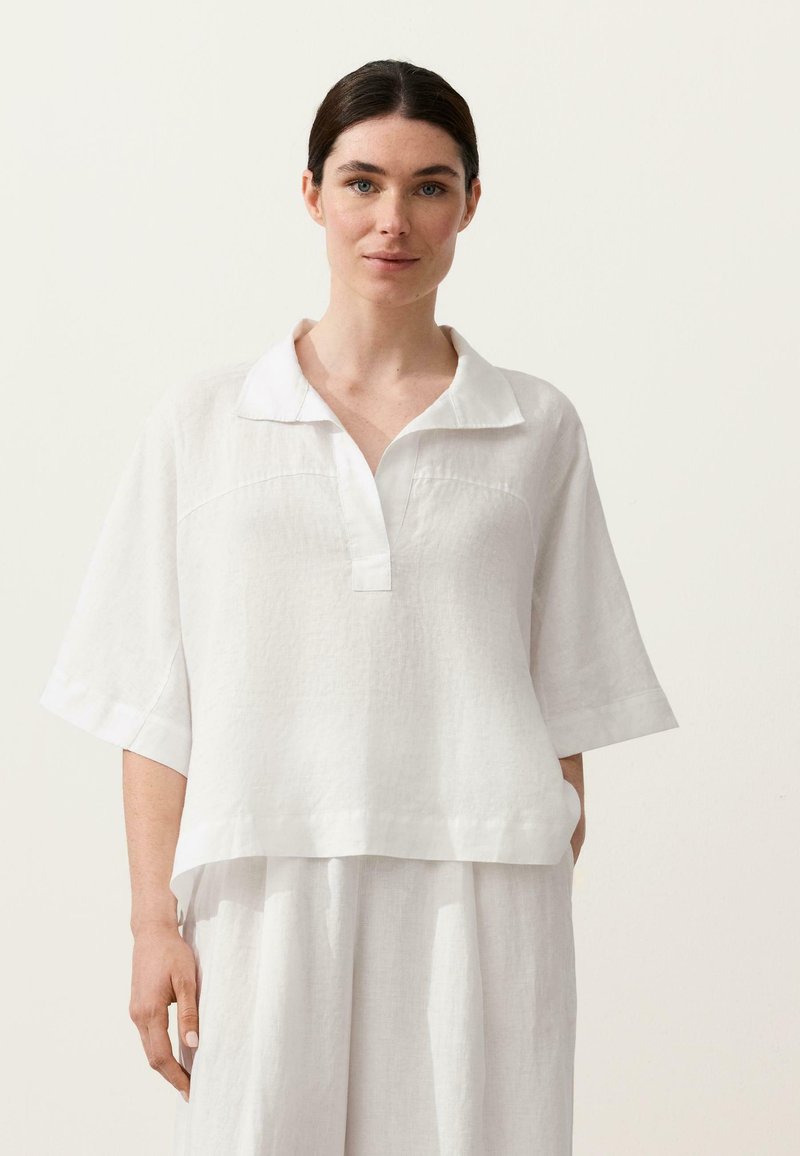 Femme aux cheveux foncés portant une chemise blanche à manches courtes amples avec col et un pantalon large assorti, sur un fond clair uni.