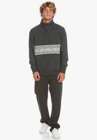 Felpa zip grigia scura con una striscia orizzontale bianca recante il logo "Quiksilver". Maniche lunghe, polsini e vita a costine. Indossata con pantaloni neri.