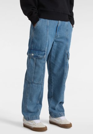 Personne portant un pantalon cargo en jean bleu avec de grandes poches latérales, des baskets blanches à semelles marron, et un haut noir à manches longues avec les mains dans les poches.
