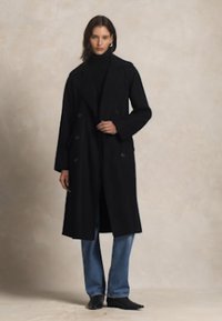Černý dvouřadý kabát z měkké látky, s volným střihem, kombinovaný s topem s vysokým výstřihem a modrými džínami.