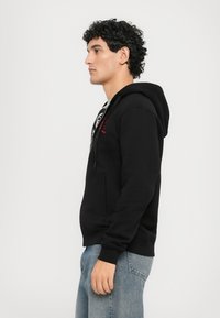 Hanorac negru cu o buzunar frontal și șnur reglabil, purtat peste un tricou gri, asortat cu blugi de culoare albastru deschis. Părul creț este vizibil.