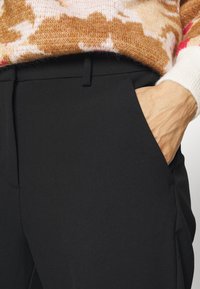 Pantaloni neri con una texture liscia, dotati di tasche laterali e una vestibilità su misura. Un maglione a fantasia con tonalità calde copre il punto vita.