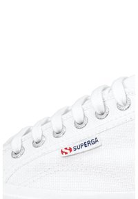 Bílé plátěné tenisky s kulatou špičkou, kovovými očky a bílými tkaničkami. Na boku se nachází modro-červené logo "SUPERGA".