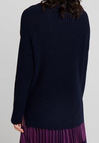 Maglione lavorato a maglia blu navy con vestibilità ampia, texture a costine e spacchi laterali all'orlo; indossato sopra una gonna plissettata viola.