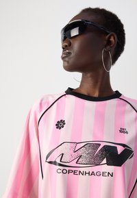 Prevelik roza striped džersy s črnimi dodatki, ki vsebuje grafični logotip in cvetlični dizajn. Model nosi temne sončne očala in obroče.