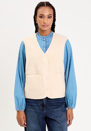 Gilet en polaire crème avec un ourlet arrondi, deux poches et des boutons-pression, porté sur une chemise en denim bleu clair à col volanté.