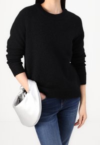 Eine Person trägt einen schwarzen Pullover und eine blaue Jeans und hält in der linken Hand eine silberne metallische Clutch-Tasche mit einem Knotenhenkel.