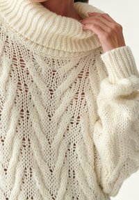 Gruba, kremowa dzianinowa sweter z szerokim prążkowanym kołnierzem, z teksturowanym wzorem w zygzak oraz luźnymi rękawami. Materiał wydaje się miękki i przytulny.