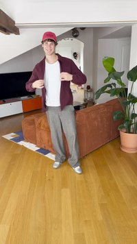 Camicia bordeaux abbottonata sopra una t-shirt bianca, pantaloni grigi a gamba larga e sneakers argento. Ambiente interno con una pianta e un divano arancione.