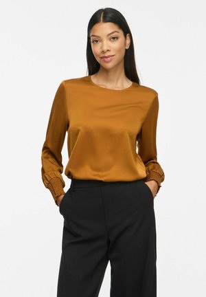 Femme aux longs cheveux foncés portant un chemisier moutarde à manches longues avec poignets smockés et un pantalon noir taille haute, mains dans les poches, souriant doucement.