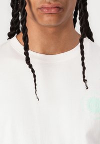 Hvid bomuld T-shirt med korte ærmer, der har en lille mintgrøn spiral grafik på venstre bryst og snoede hårtråde i fokus.