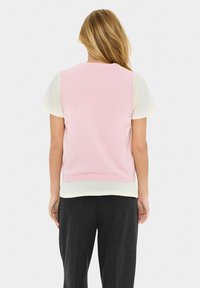 Un pull sans manches rose superposé sur une chemise crème claire, avec un pantalon gris foncé. La texture douce et les bords côtelés mettent en valeur le design.