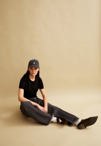 Zwarte t-shirt, donkergrijze wijde jeans en een grijze pet met een wit logo. Bruine schoenen met spitse neuzen en een accent van een bandje. Beige achtergrond.